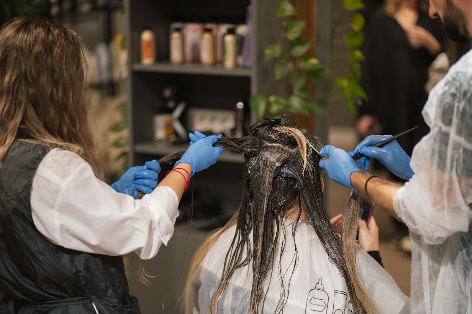 Two stylists in gloves sectioning a guest's damp hair and brushing product through it at a salon station