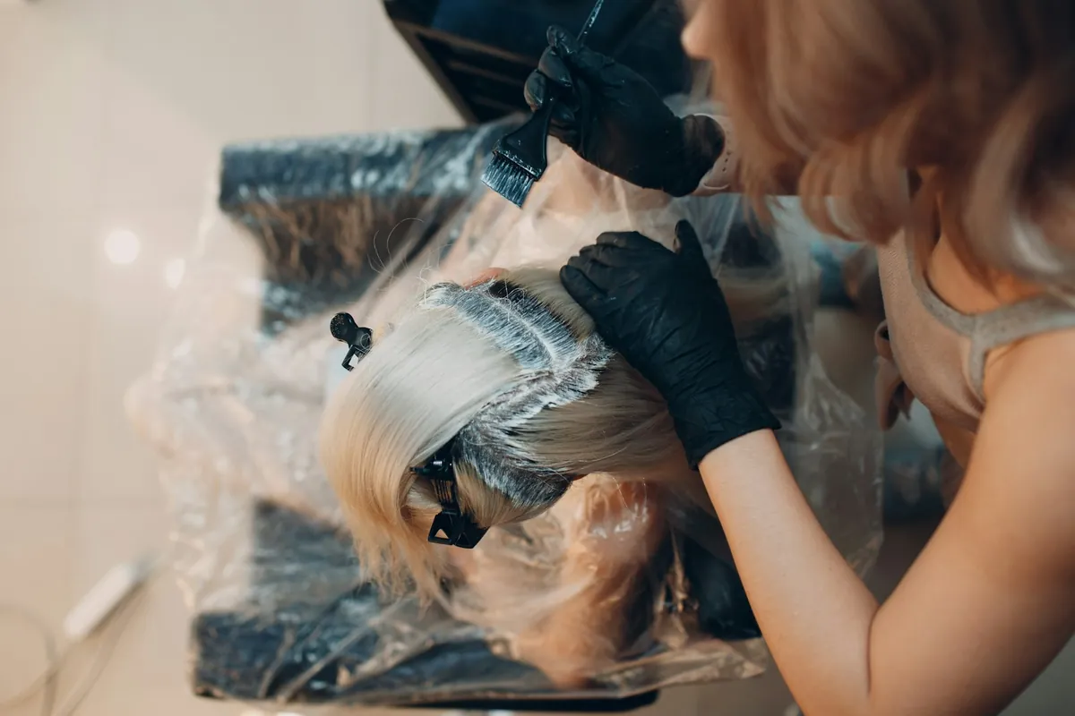 Salon Bastille style color service: a gloved colorist applying color along the root line of a guest's silver blonde hair at a salon station